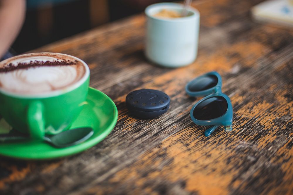 A blurry shot of a latte in a green cup next to small sunglasses on worn-out wooden surface. Original public domain image…