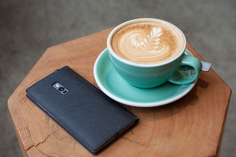 A smartphone on a wooden surface next to a cup of coffee with latte art. Original public domain image from Wikimedia Commons