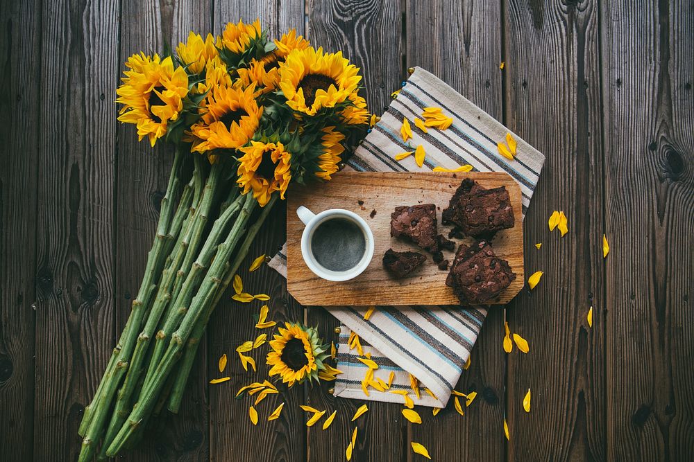 A bouquet of sunflowers, striped placemat, wooden board, cup of coffee, and brownies on a table. Original public domain…