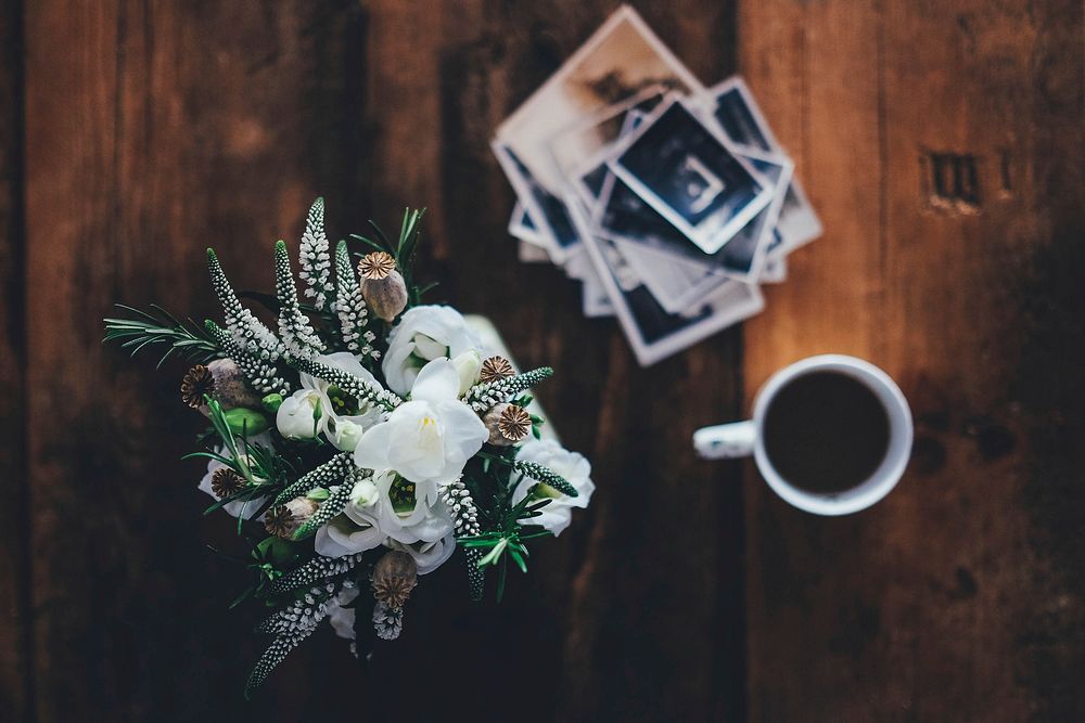 An overhead shot of a bouquet of flowers next to a cup of coffee and a stack of photographs. Original public domain image…