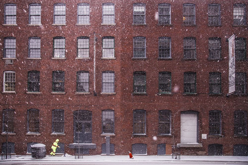 A person bundled in reflective yellow clothing shovels a sidewalk in front of a large red brick building during a snowfall.…