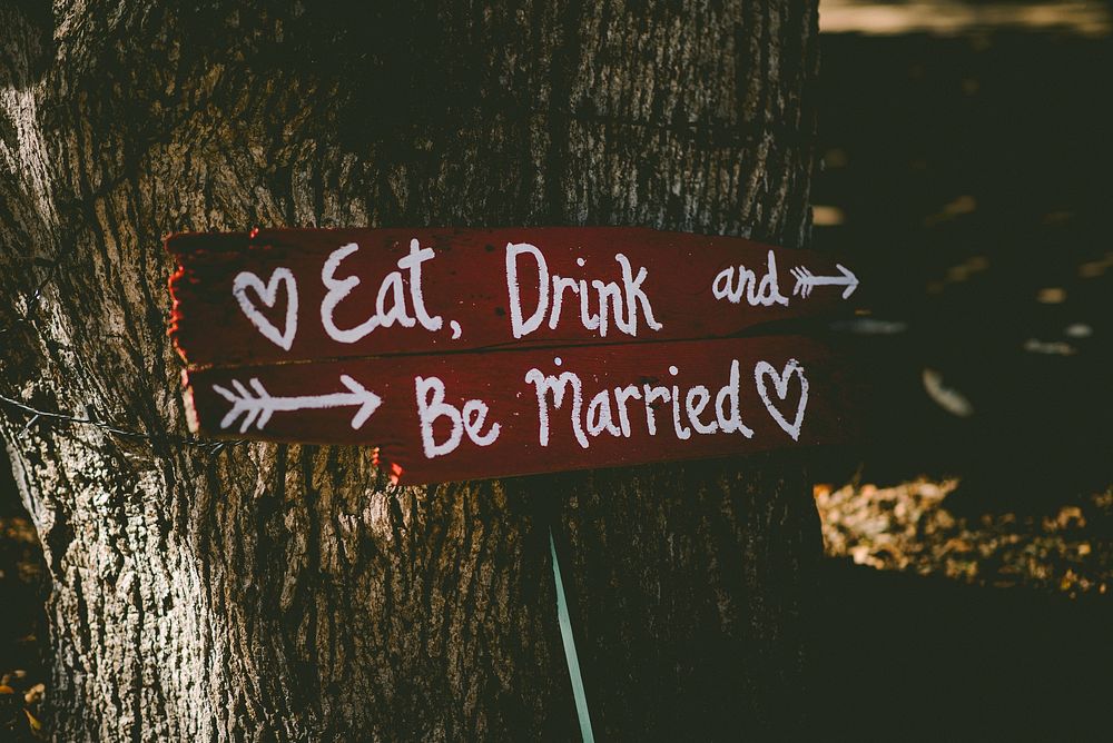 Red sign says "Eat, drink and be married" in white writing in front of a tree trunk. Original public domain image from…