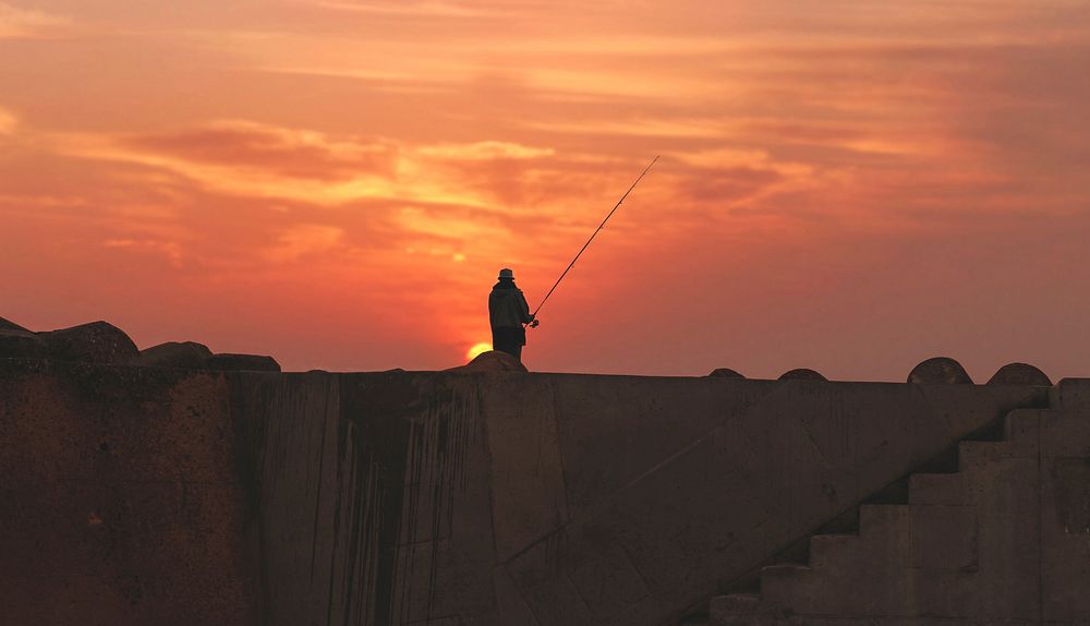 A silhouette of a fisherman standing atop a wall in Asilah, Morocco during sunset. Original public domain image from…