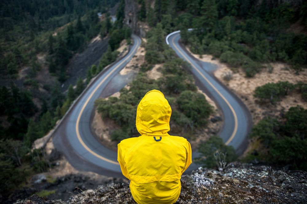 A person in a yellow jacket with a hood sitting on a rock over a U-turn in a road. Original public domain image from…