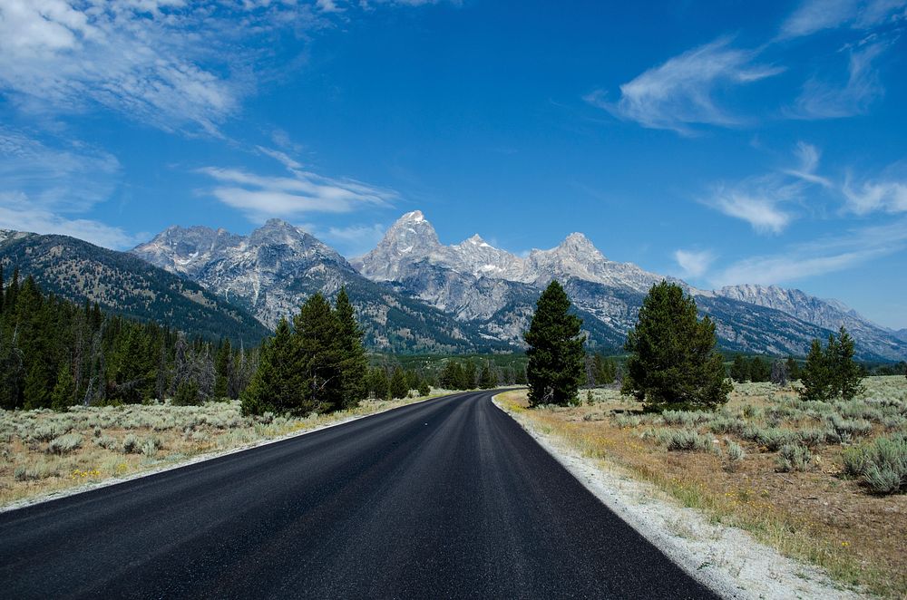 A winding road with green pine trees and a mountain range in the distance at Grand Teton. Original public domain image from…