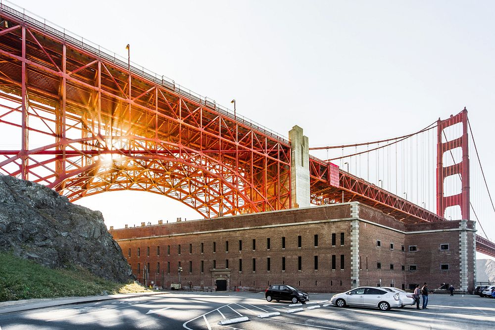 San Francisco Golden Gate Bridge at sunset suspended over a brown building, cars and people. Original public domain image…