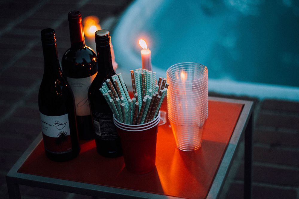 A tabletop containing wine, straws, candles with flames and plastic cups by a pool in redding. Original public domain image…