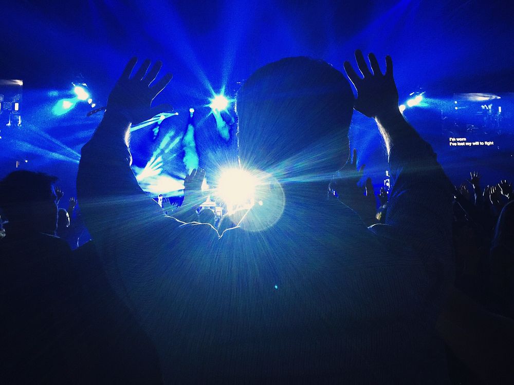 The silhouette of a person at a concert throwing their hands up in the blue light. Original public domain image from…