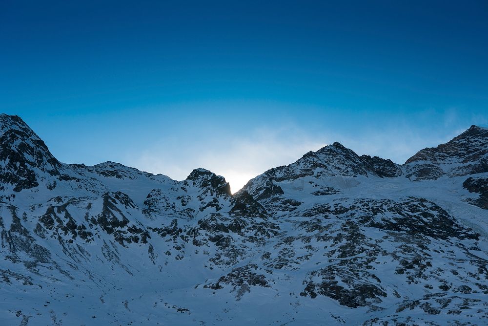 Sun hiding behind shaded mountain slopes covered with a snow blanket. Original public domain image from Wikimedia Commons