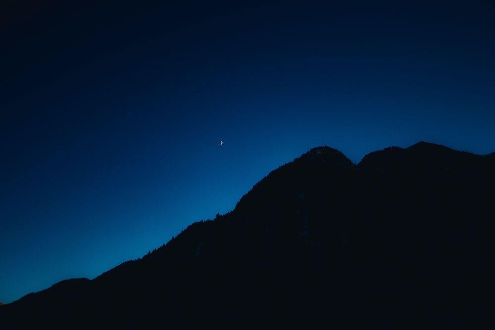 A silhouette of a wooded mountain under an evening sky with a crescent moon. Original public domain image from Wikimedia…