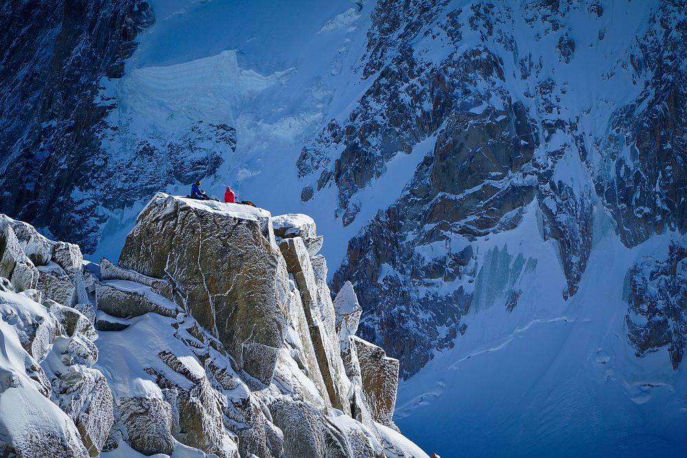 Two people wearing winter jackets sitting on a cliff on the side of a snowy mountain during winter in Chamonix. Original…