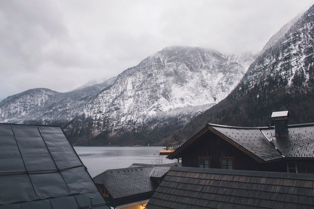 Rooftop view of houses and snow covered mountains surrounding it with water in Hallstatt.. Original public domain image from…