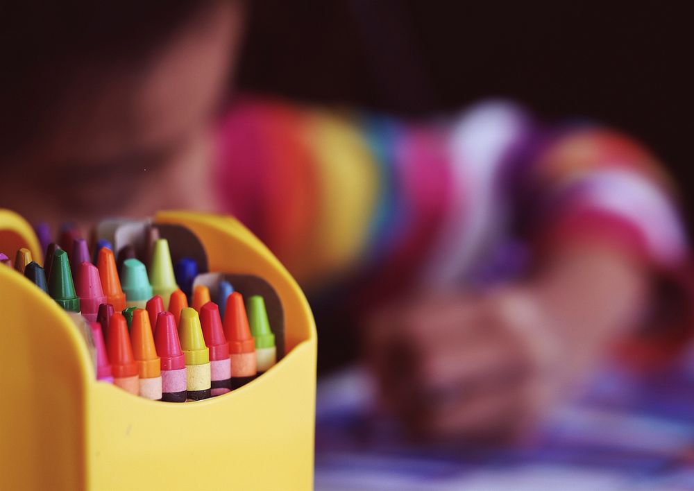 A child, wearing a colorful shirt, uses crayons to color a picture.. Original public domain image from Wikimedia Commons
