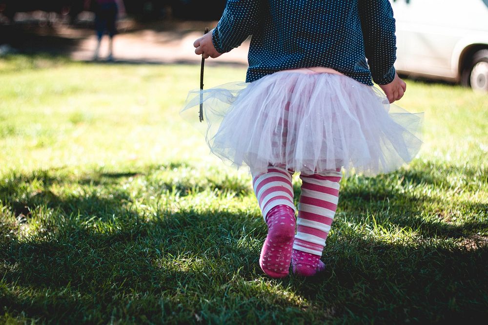 Female child wearing a tutu with stripe leggings walks on grass towards sunlight holding a stick. Original public domain…