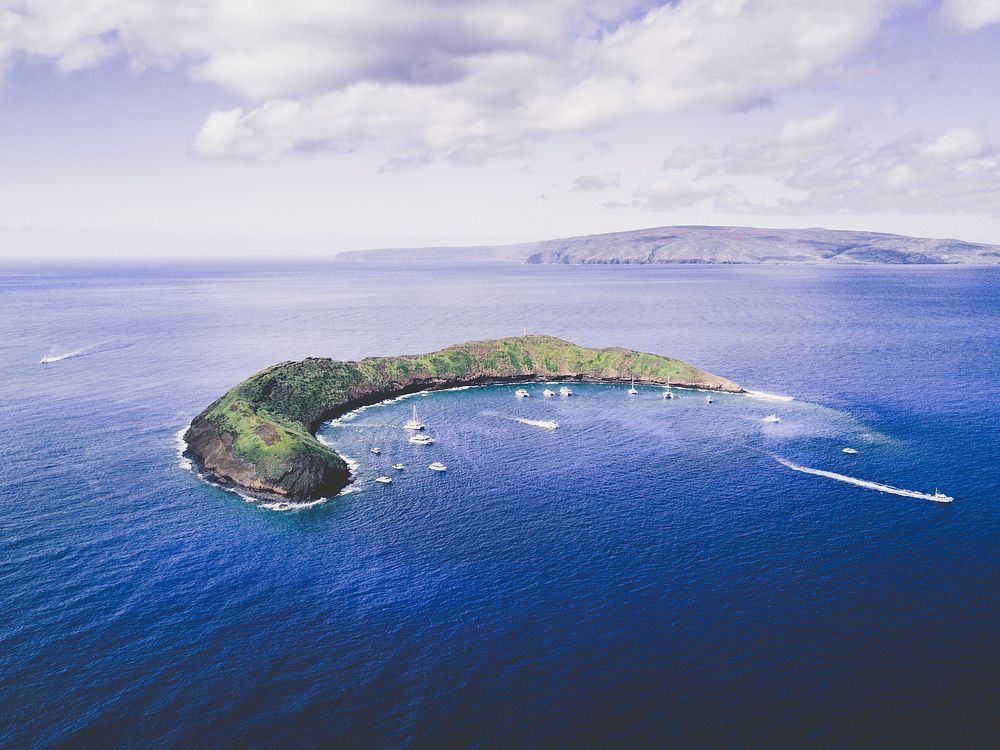 A crescent moon shaped island in the middle of a bright blue ocean at Molokini Crater. Original public domain image from…