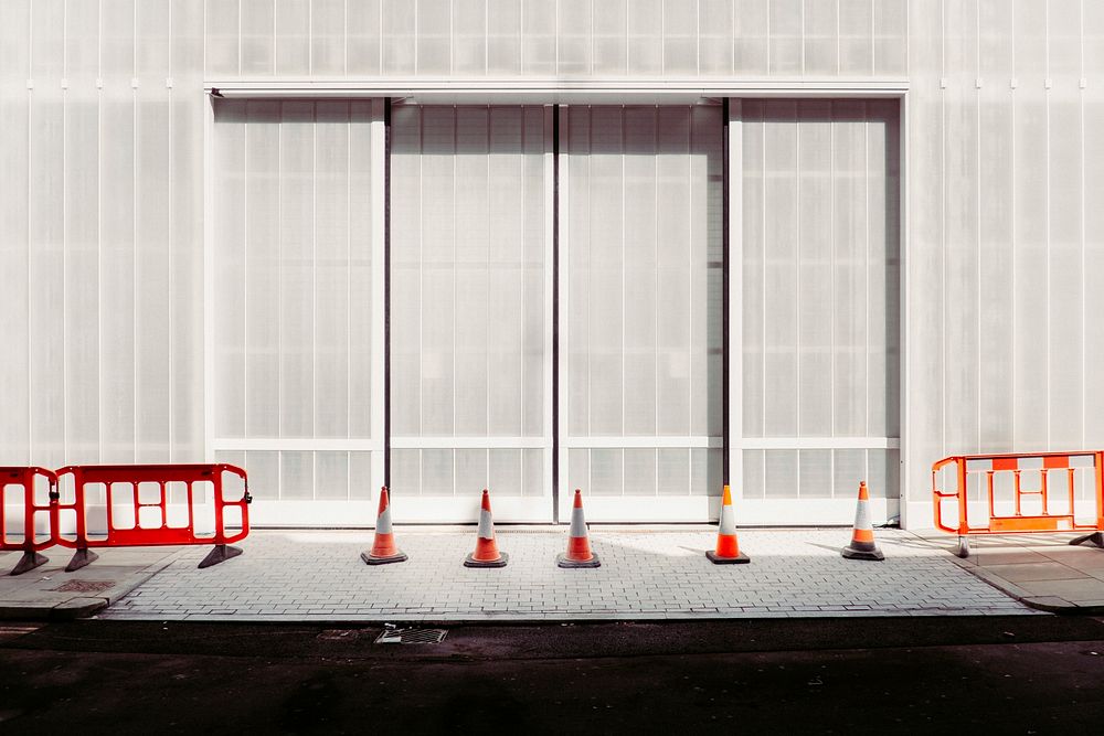 London sidewalk with orange construction cones and guard rails in front of a gray building. Original public domain image…