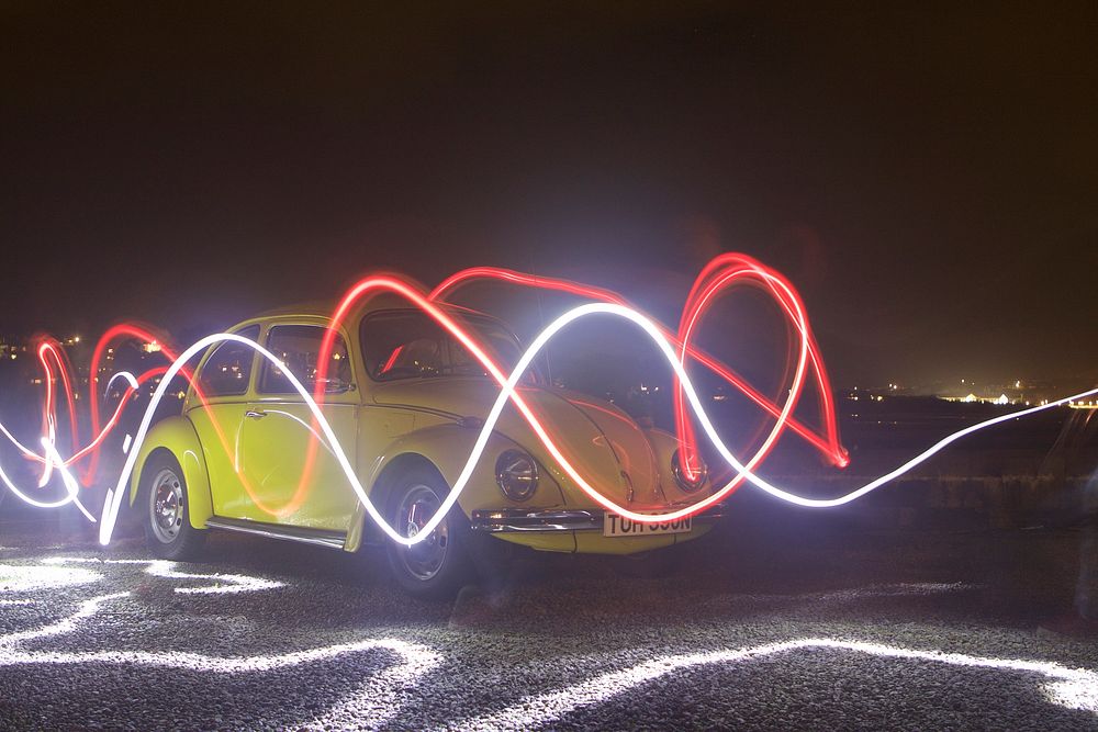 Red and white light trails drawn in the air in front of the yellow vintage Volkswagen beetle at night. Original public…