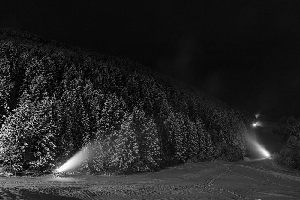 A dark black and white shot of a forest with snow-covered trees and spotlights. Original public domain image from Wikimedia…