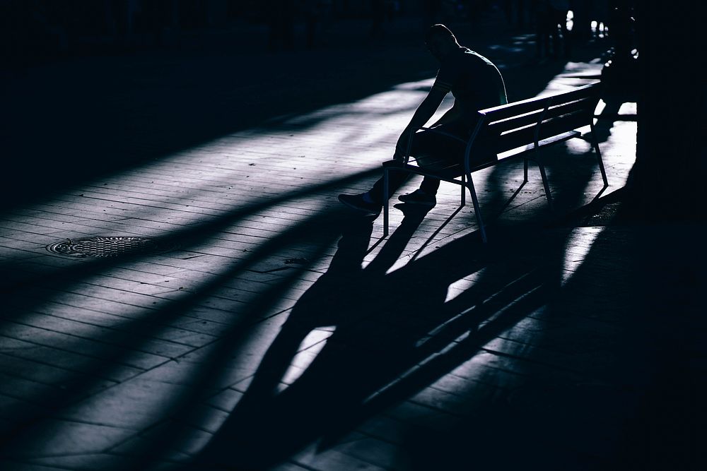 A dim shot of a person sitting on a bench and casting a long shadow. Original public domain image from Wikimedia Commons