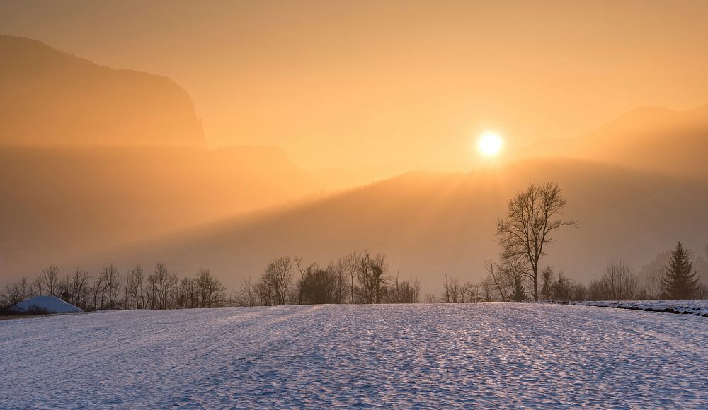 Golden rays of the sunset hit a barren snowy valley with mountains and trees in the distance. Original public domain image…