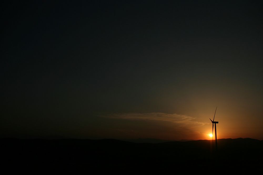 The sunset shines a bright light upon a Pikrolimni wind turbine standing in the darkness of the night. Original public…