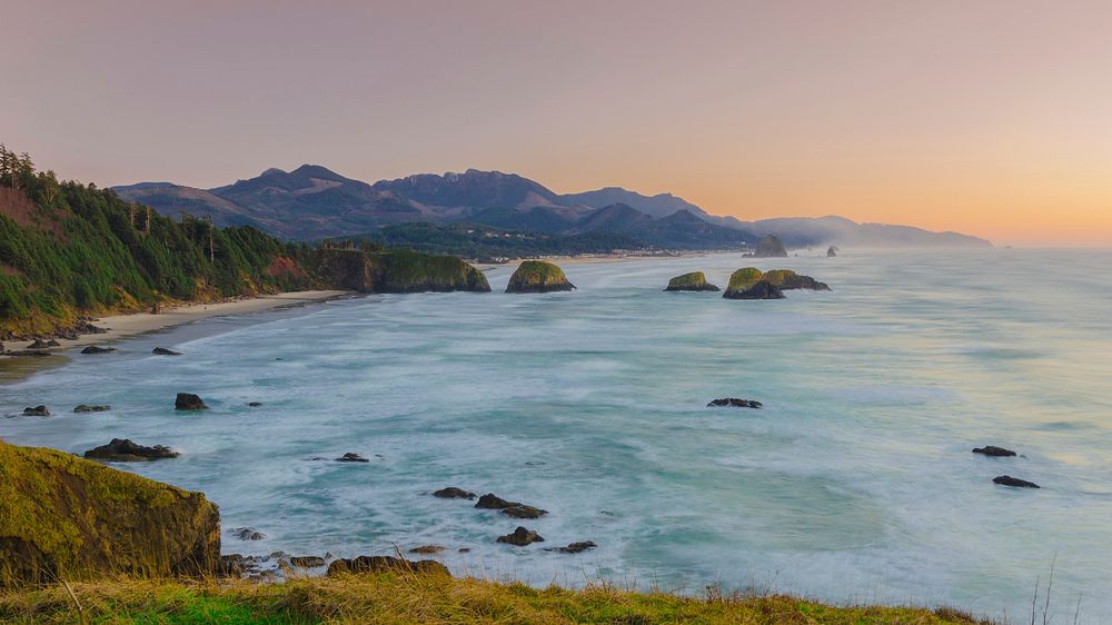 Ocean washing on a sandy beach with green cliffs in Ecola State Park at sunset. Original public domain image from Wikimedia…