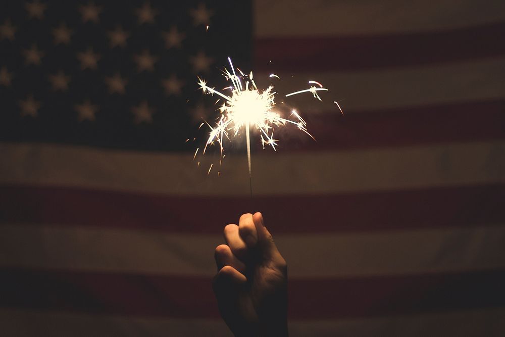 A person's hand holding a sparkler in front of an American Flag. Original public domain image from Wikimedia Commons
