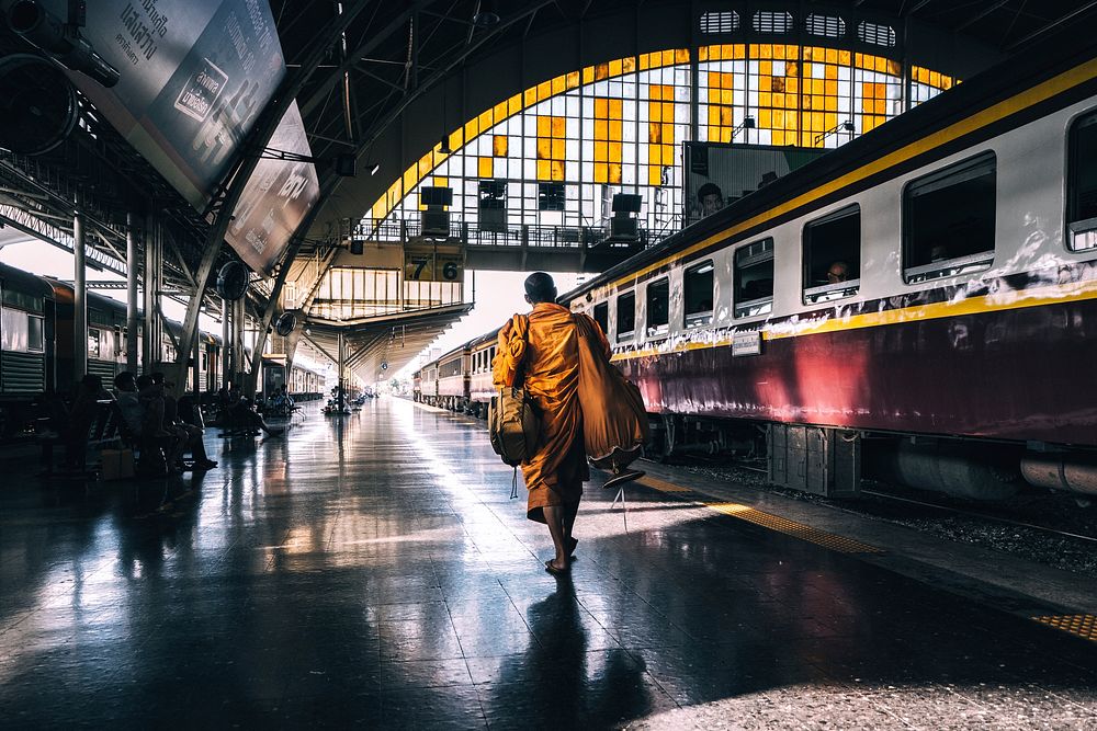 A monk in orange robes walking across a railway platform in Bangkok. Original public domain image from Wikimedia Commons