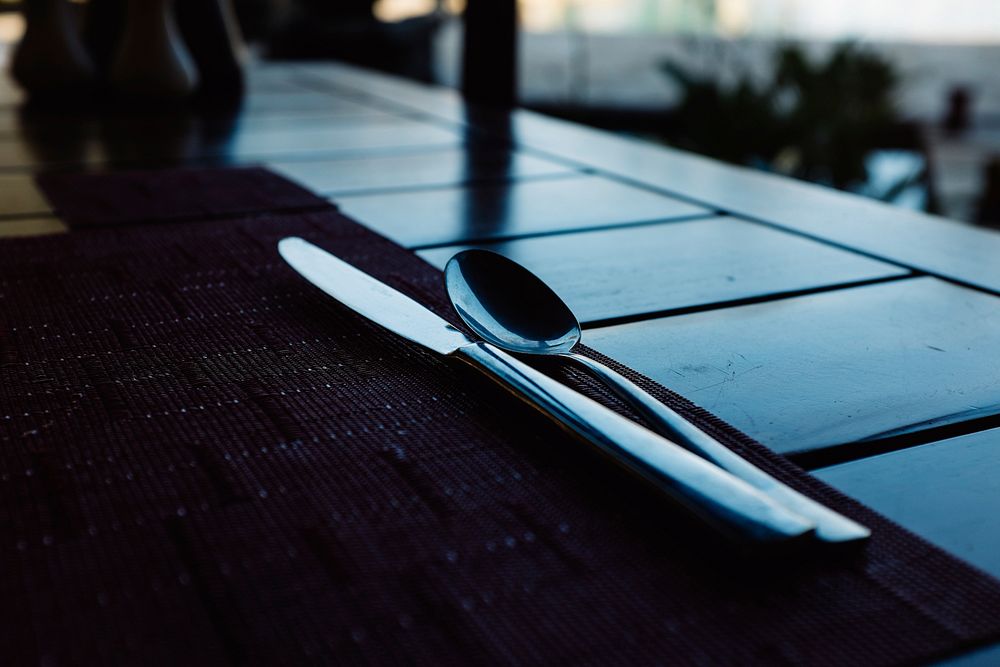 Macro of shiny knife and spoon on a placemat near a window on a slatted table. Original public domain image from Wikimedia…