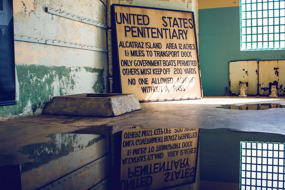 Puddle in an abandoned jail cell reflects old yellow sign for the United States Penitentiary.. Original public domain image…