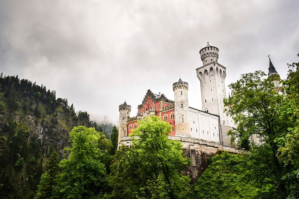 The fortifications of Neuschwanstein Castle rising up above green trees. Original public domain image from Wikimedia Commons