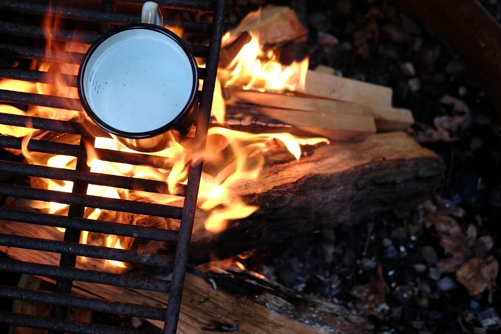 A tin cup of water on a grill above a campfire made of wooden logs in a fire pit. Original public domain image from…