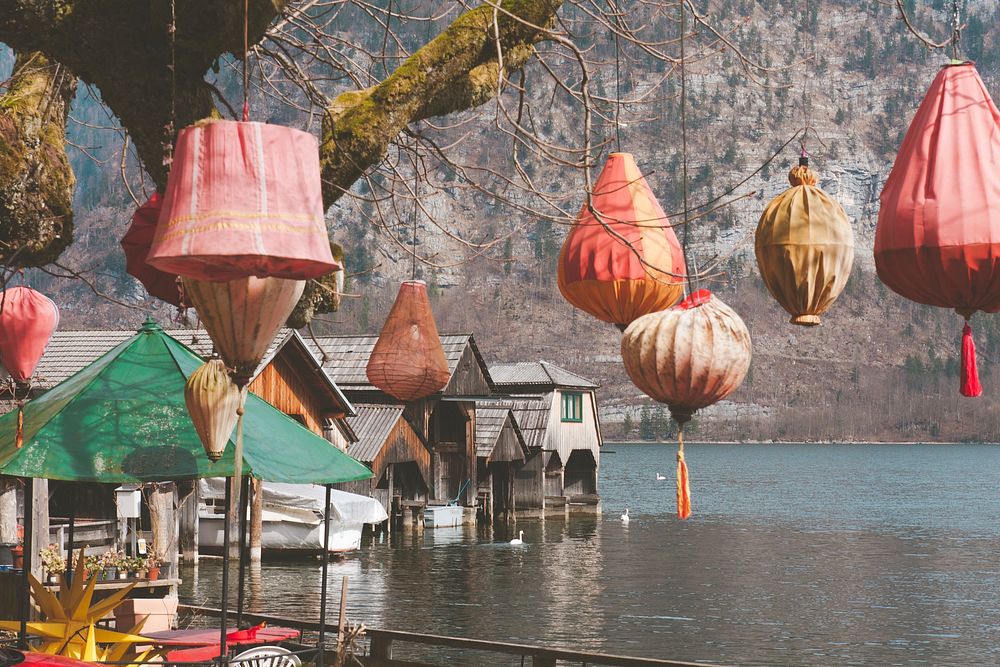 Wooden Buildings Sitting on Lake Hallstatt framed with red and orange lanterns hanging from a tree. Original public domain…