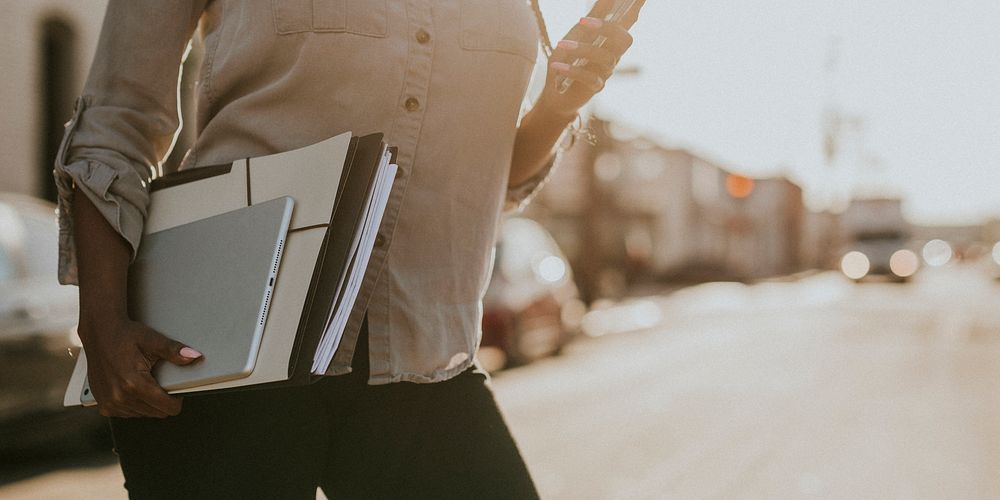 Black woman with file folders crossing a street while using her phone