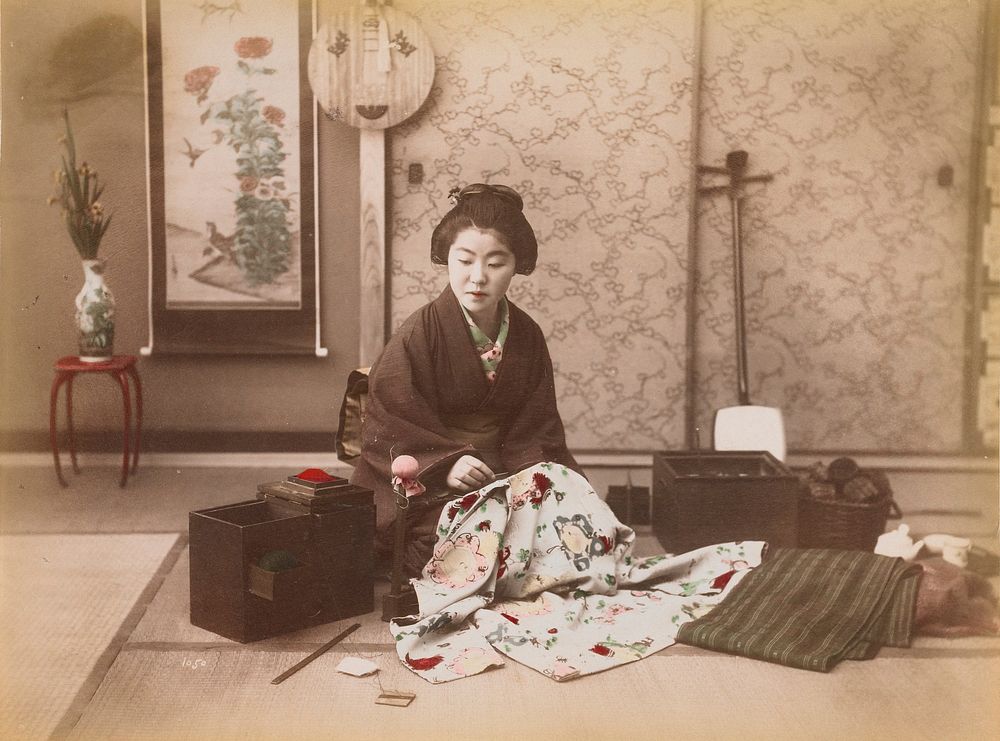 young woman wearing traditional Japanese garments seated in an interior, sewing, with sewing box to her right and fabrics on…