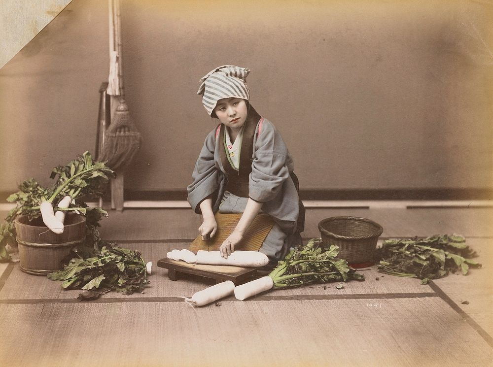 young woman wearing a grey kimono and blue and white striped head scarf, cutting up daikon radishes. Original from the…