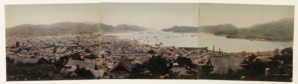 panoramic view of densely packed rooftops and trees along a bay surrounded by mountains, and filled with boats. Original…