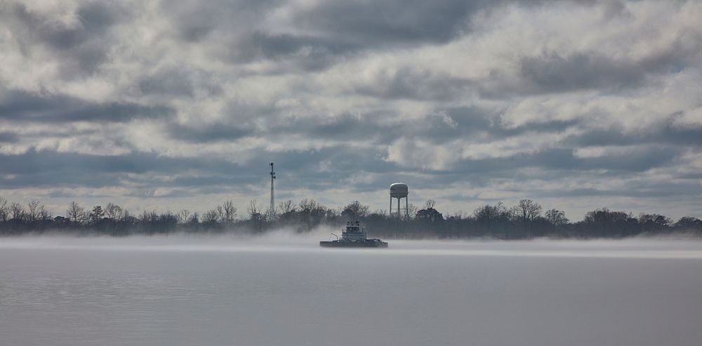                         A tugboat pushes barges loaded with coal on the misty Mississippi River as it winds through rural…