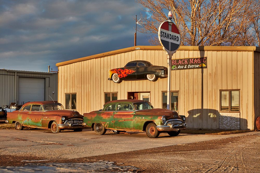                         Two automobiles that may (or may not) be beyond restoration sit outside the Black Magic Rod & Custom…