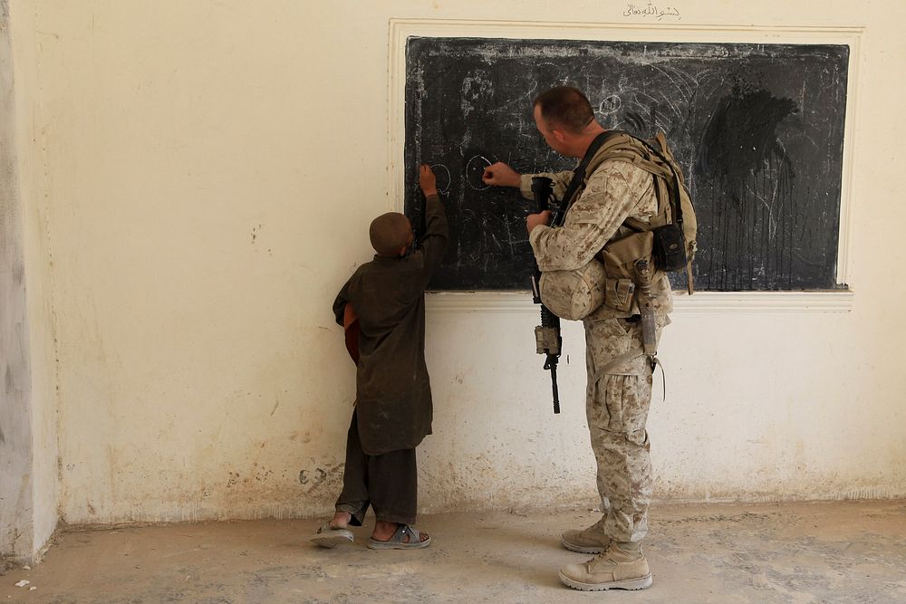 U.S. Marine Corps Maj. Nicholas Martz, from 1st Battalion, 5th Marine Regiment, writes on a chalkboard with an Afghan boy…