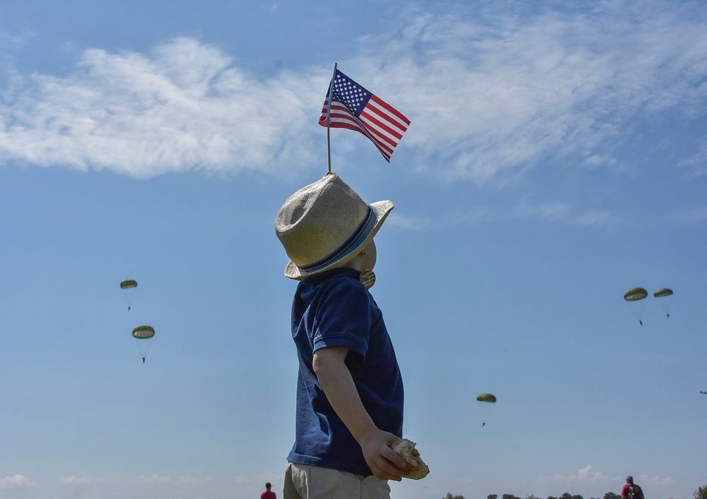 A young boy dressed in a Fedora and waving an American flag welcomes paratroopers as they jump into Sainte Mere Eglise…