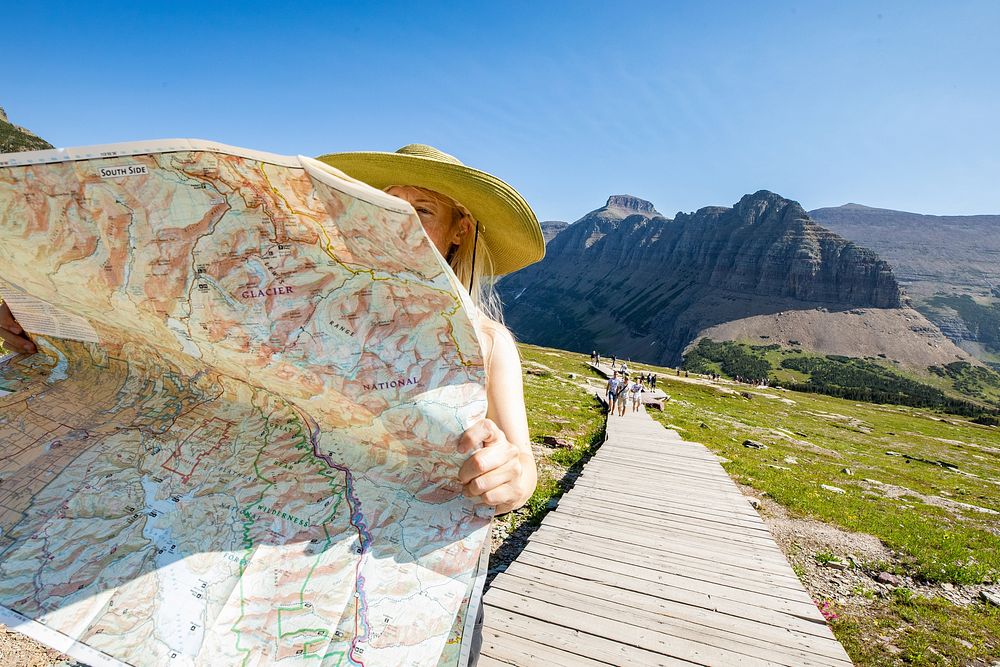 A park visitor stands on a boardwalk in the mountain meadow of Logan Pass and reads a park map with a sunhat on. Original…