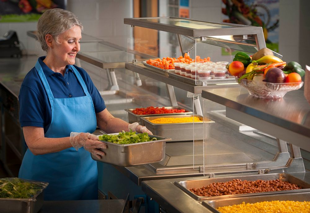 School Nutrition Professional preparing the school lunch serving line with vegetables for a burrito bar. Original public…