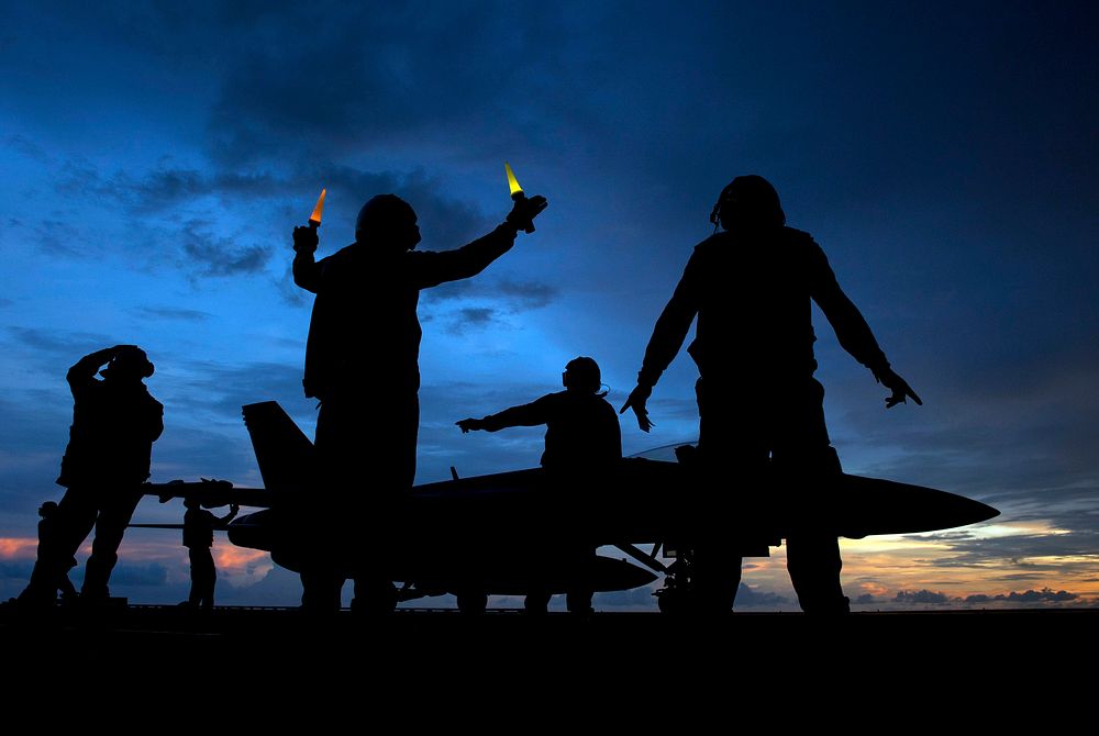 A U.S. Navy aircraft director signals as aviation ordnancemen unload ordnance from an F/A-18E Super Hornet aircraft aboard…