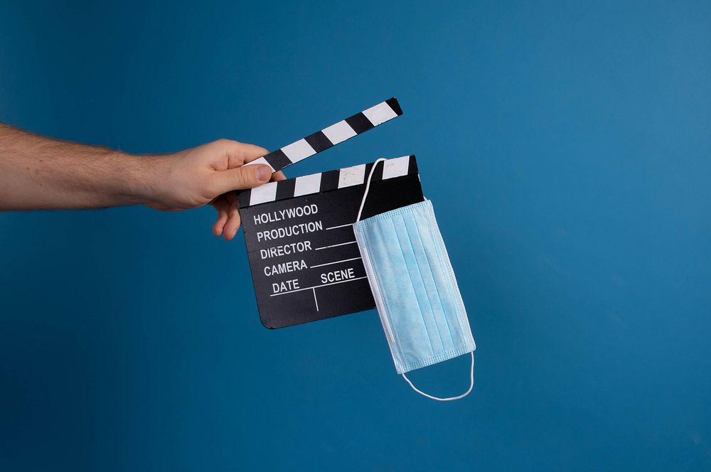 Hand holding movie clapper and medical face mask on blue background 