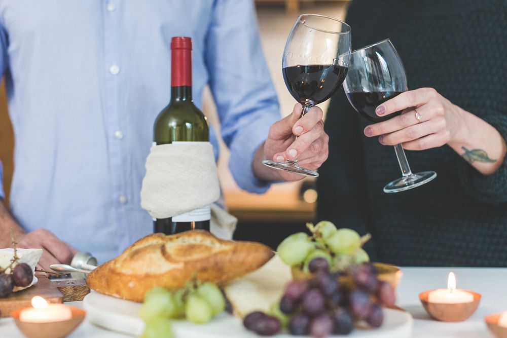 Couple toasting glasses of red wine with snacks set out on table.