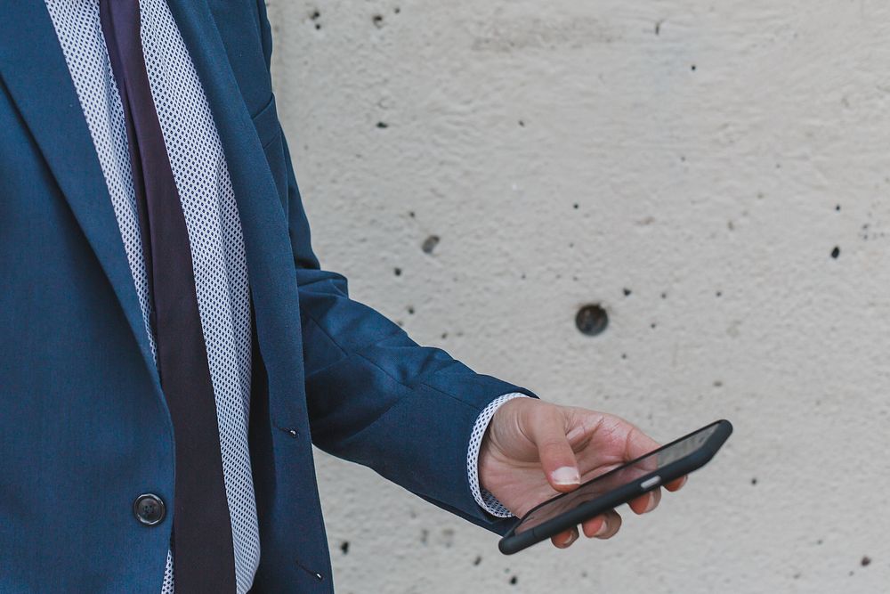 Close up of man holding a smart phone standing in front of cement wall.
