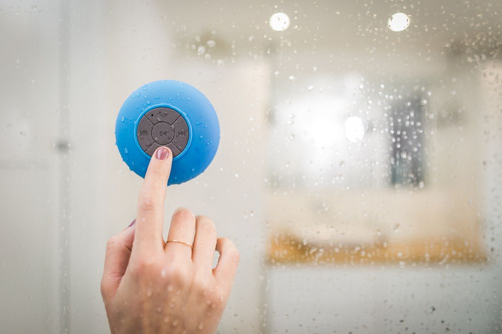 Womans finger pressing option button on blue waterproof speaker in shower.