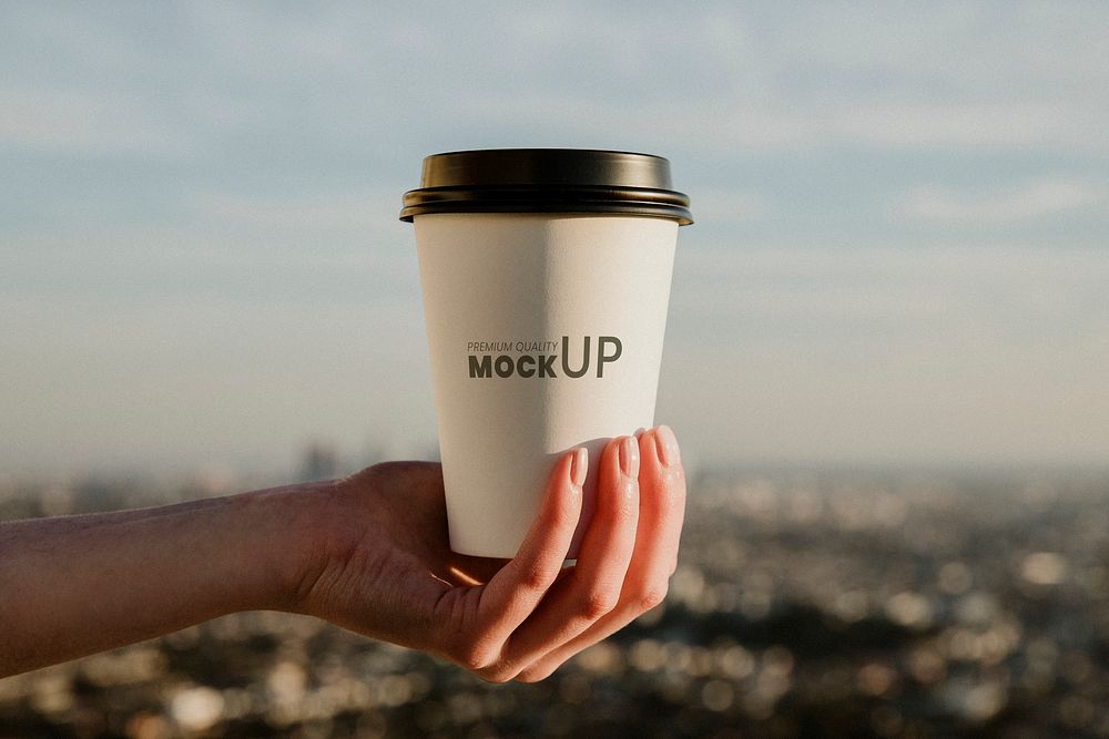 Hand holding a takeout coffee cup with urban view mockup