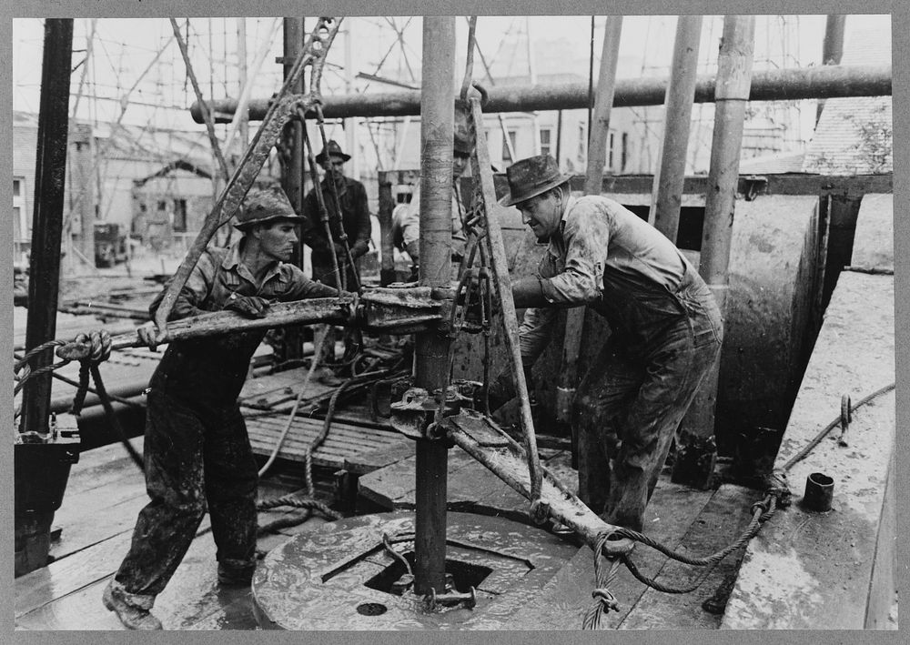 Oil roughnecks screwing one piece of drill pipe to another piece by means of a heavy pipe wrench, Kilgore, Texas by Russell…